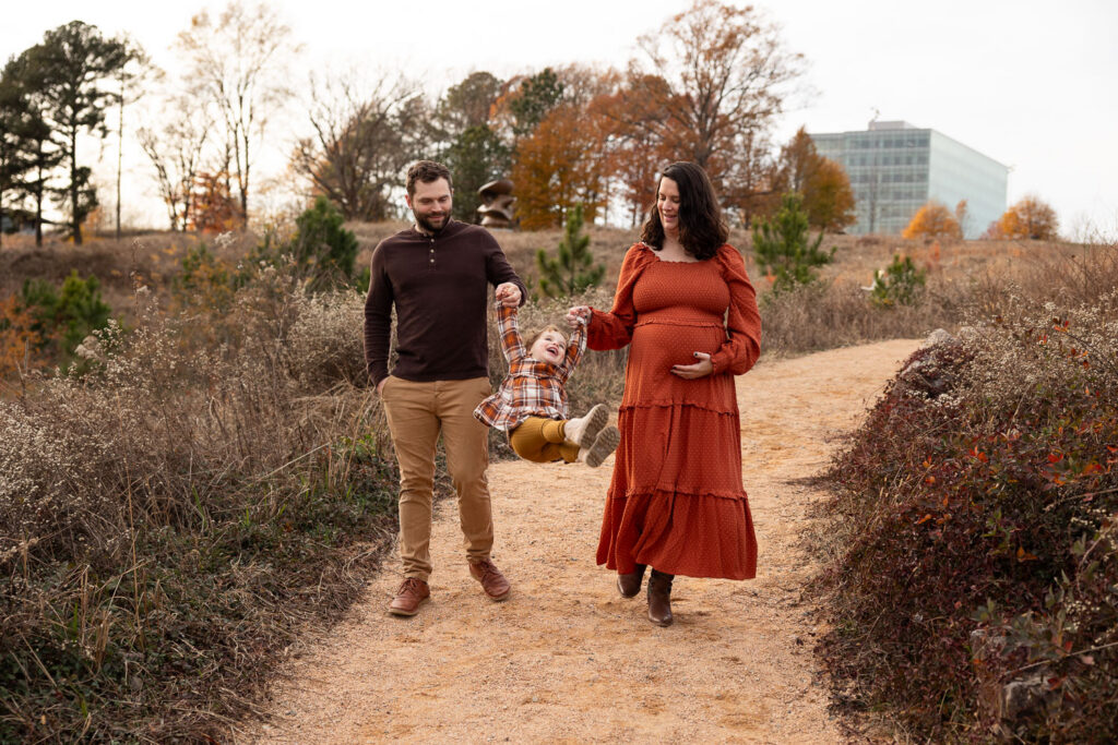 Raleigh family of three maternity photos at NCMA, parents walking toward camera while swinging daughter between them