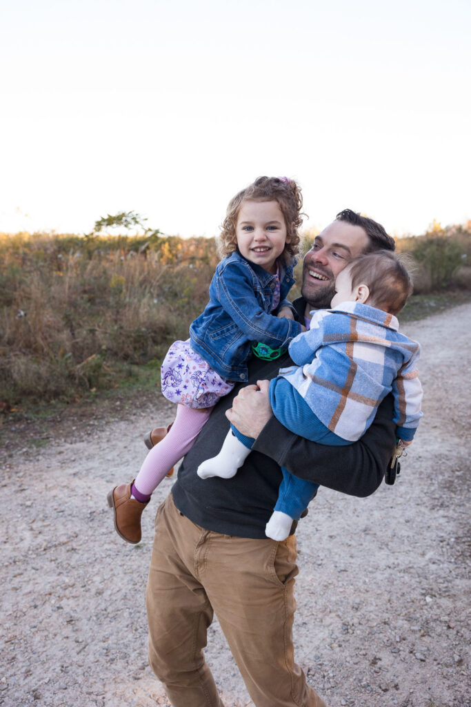 Dad leans back while holding toddler daughter and 9-month old baby son during family photo session in Raleigh