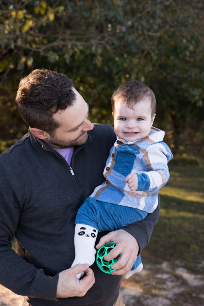 9-month-old baby boy smiles at camera and is held by his dad during baby photo session at Prairie Ridge Ecostation