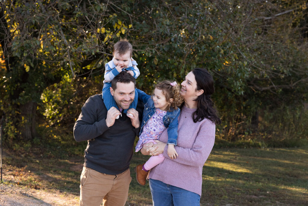 Baby's first year photo collection session for 9-month old baby at Prairie Ridge Ecostation in Raleigh, family of four stands together with mom holding daughter and baby boy on dad's shoulders
