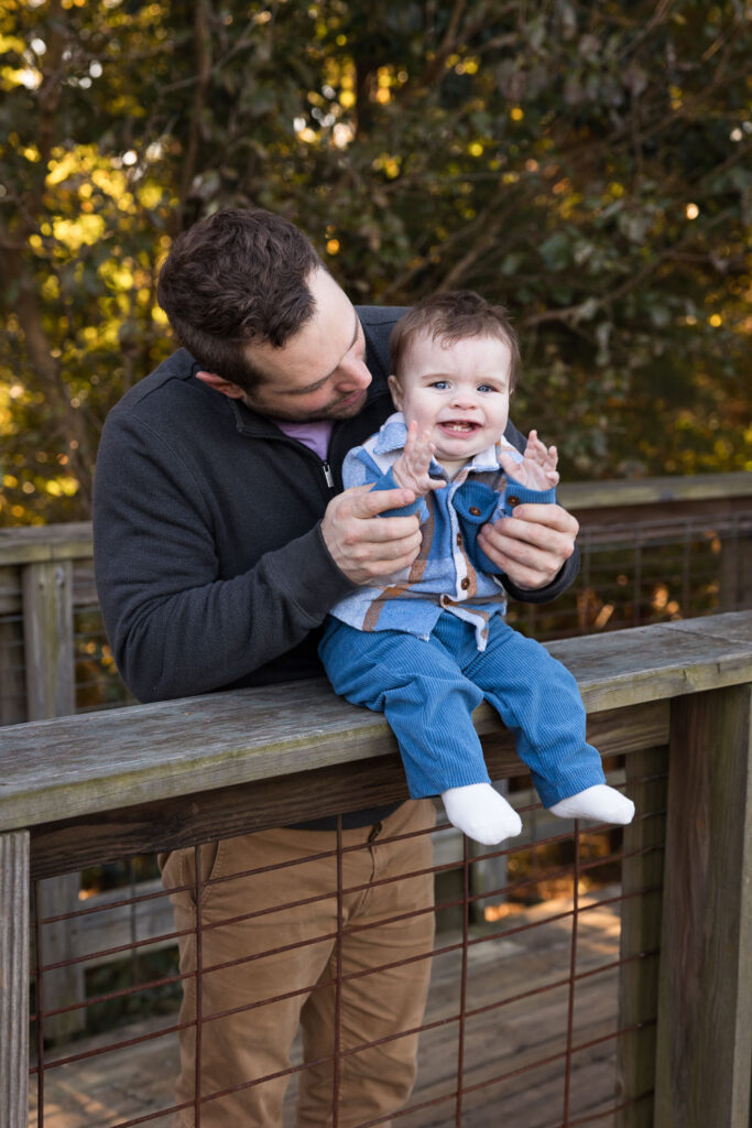 Prairie Ridge Ecostation family photo session with dad and baby, dad holds baby while baby claps and smiles while sitting on railing