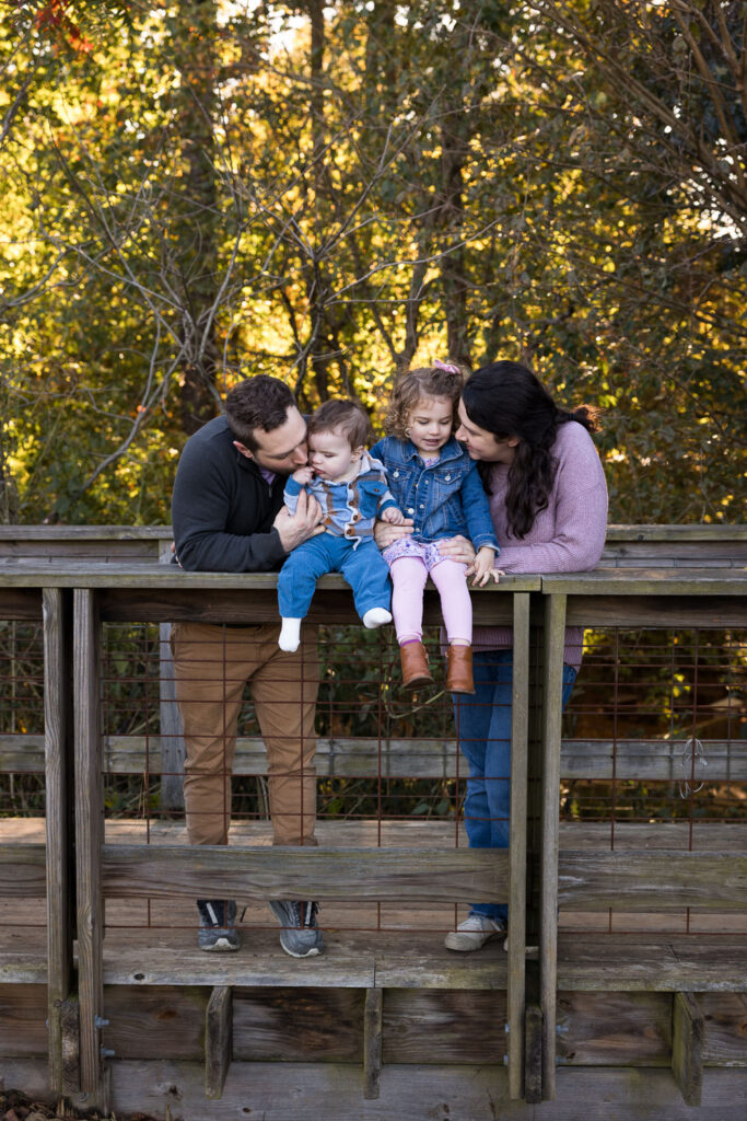 Prairie Ridge Ecostation baby milestone photo session, baby and toddler sit on wooden railing while mom and dad snuggle in and support them