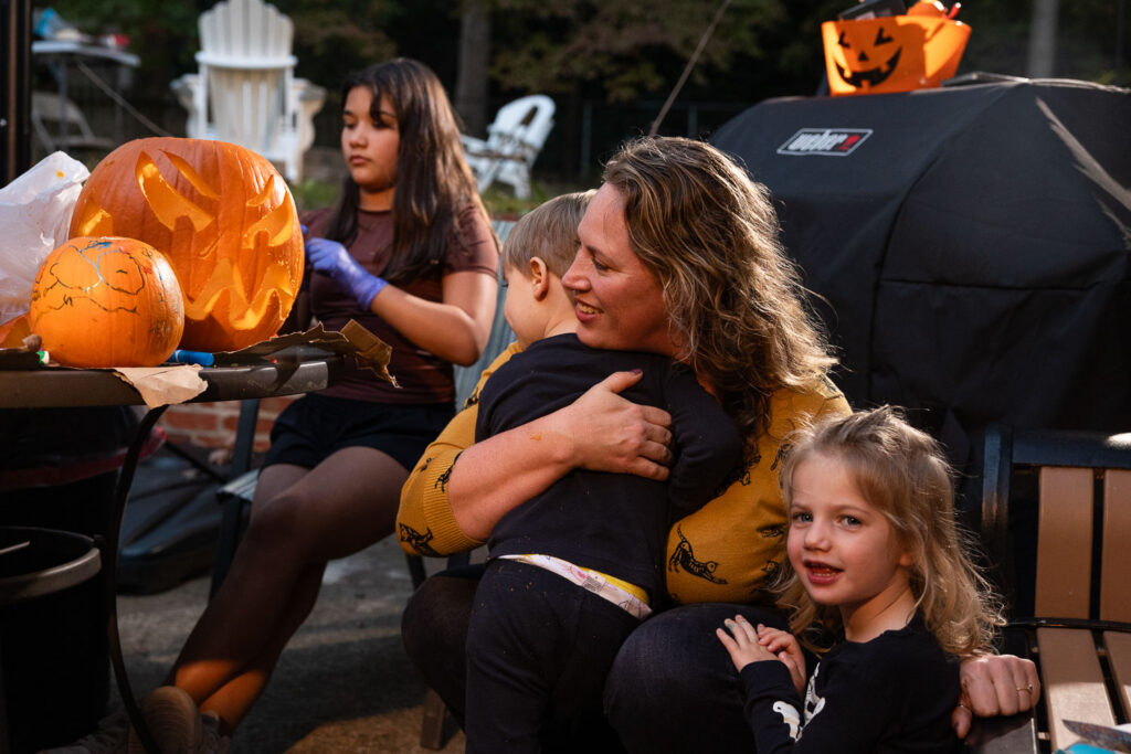 Candid fall photo session carving pumpkins, mom hugs her twin toddlers next to the pumpkin carving table in a candid moment of love