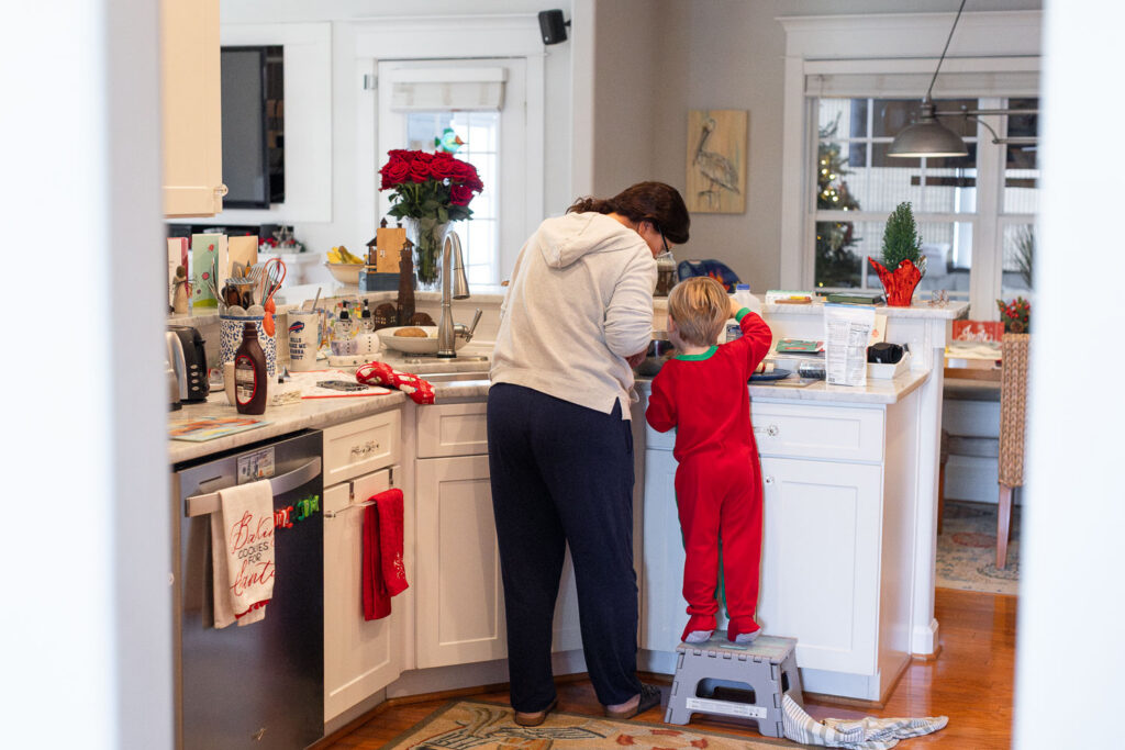Raleigh holiday family photos capturing holiday traditions. A grandmother stands next to her young grandson who is on a stool in the kitchen and they are mixing dough to bake.