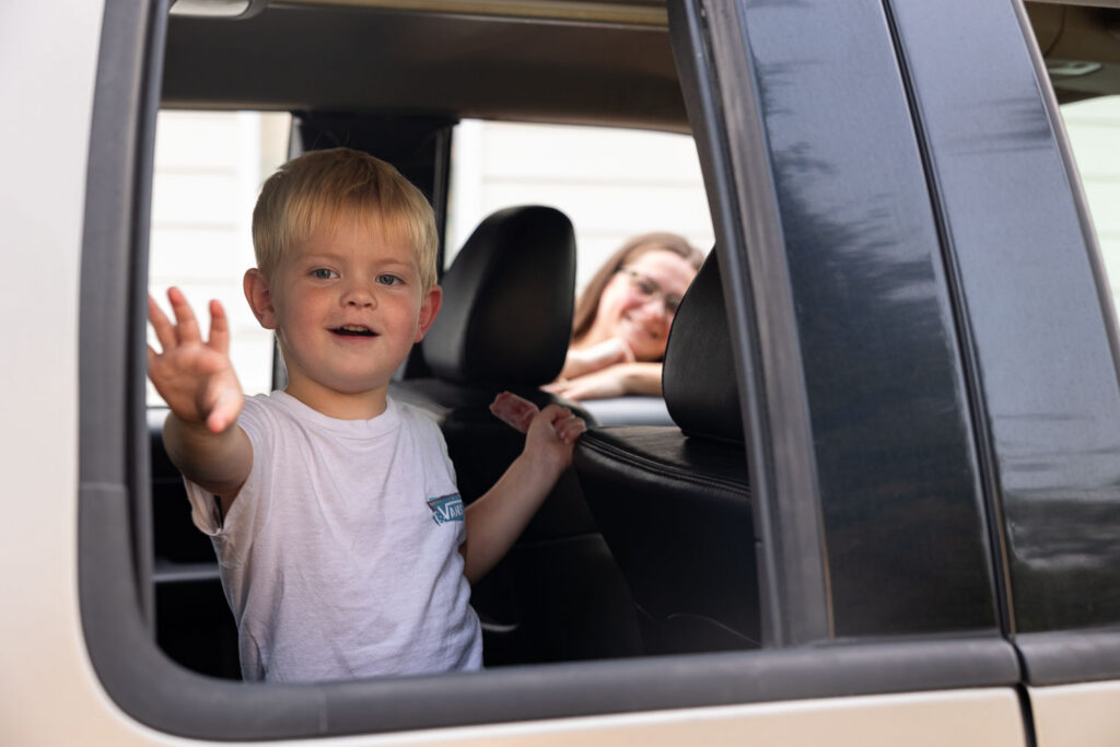 Young boy waving through the open car window while his mom smiles from the front seat.