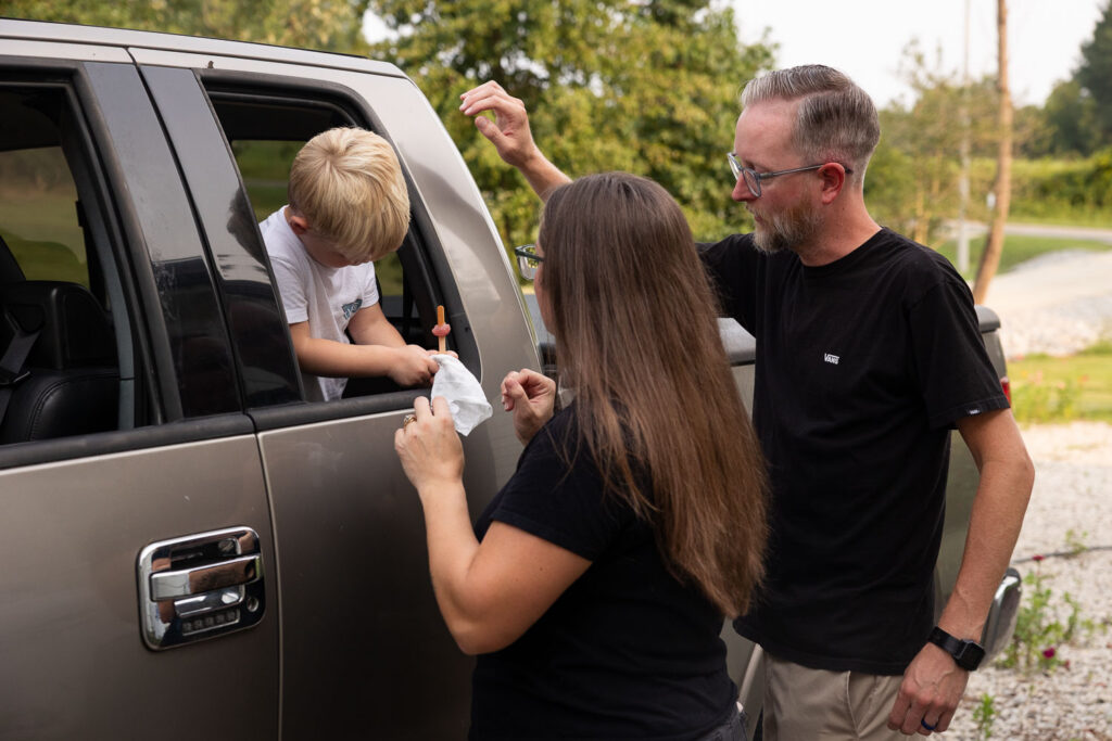 Parents chatting with their young son through the open car window as he cleans off his own hands with a wipe.