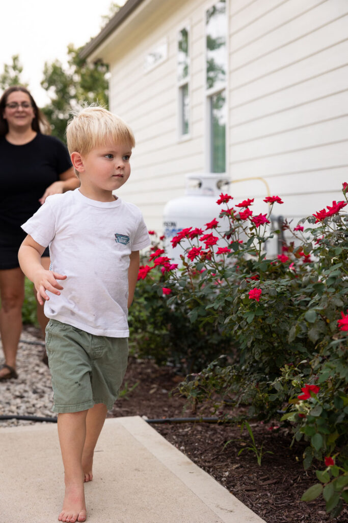 Young boy walking along the sidewalk beside red flowers outside his family’s home.