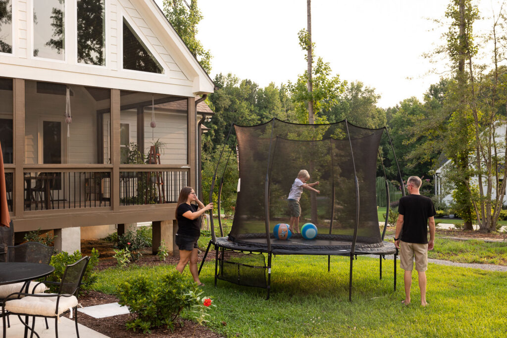 Backyard view of a family home with a trampoline where parents and their young child play together.
