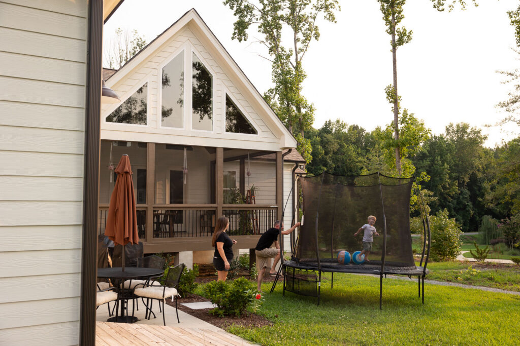 Wide view of a family’s backyard, home, and trampoline on a summer evening as the dad begins to climb onto the trampoline.