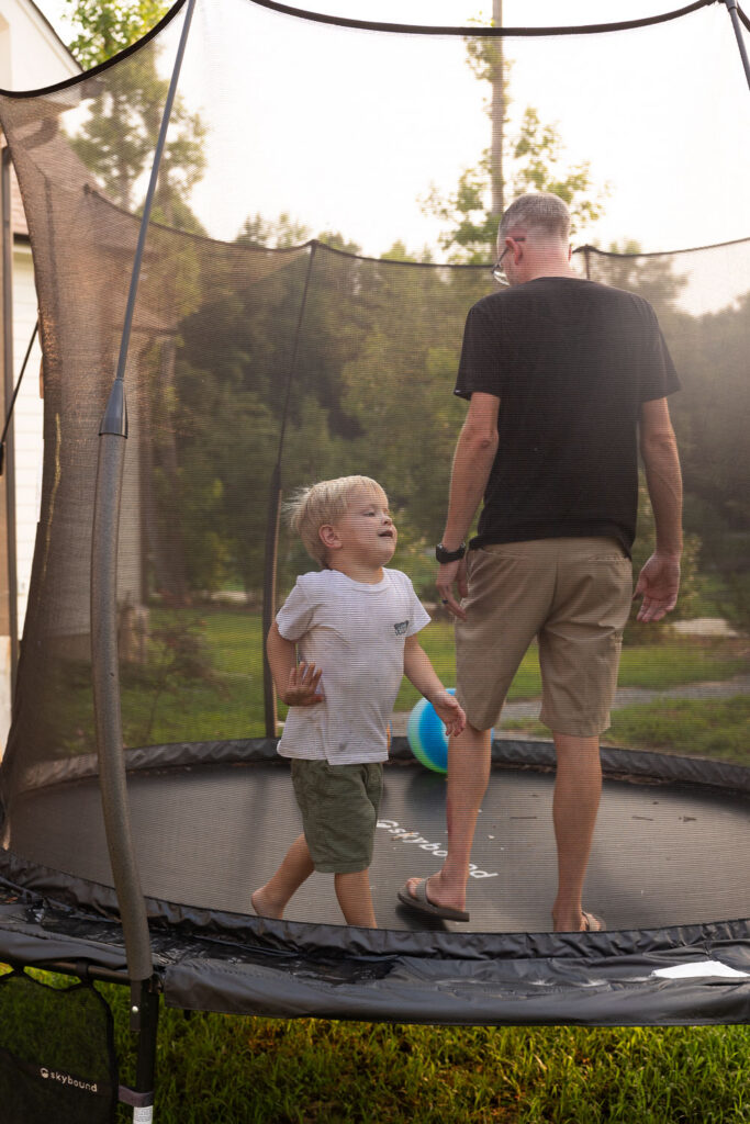 Dad standing on the backyard trampoline beside his young son.
