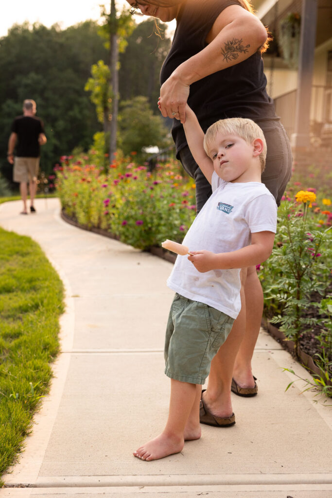 Toddler son holding his mom's hand and leaning back playfully beside the flower garden outside their home.
