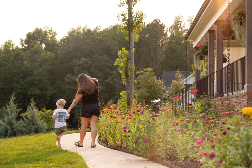 Mom and young son walking along the front sidewalk in front of the house beside a colorful flower garden.