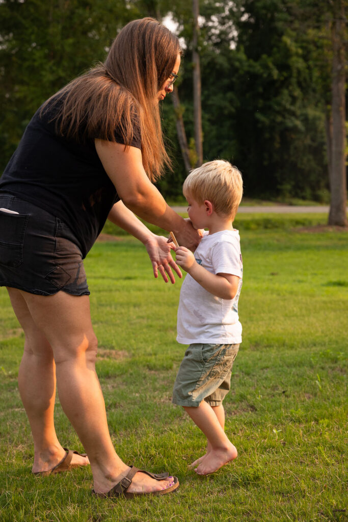 Mom and young son stand together on the lawn as the toddler uses his mom's hand to wipe his chin.