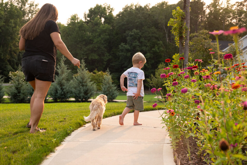 Young boy and family dog walking ahead of mom along the path beside their home.