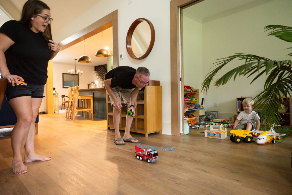 Mom and dad watching and interacting with their young son as a firetruck toy zooms across the floor.