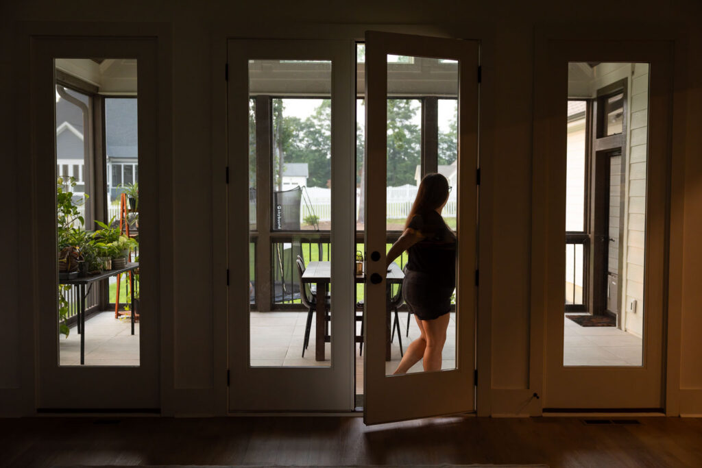 Silhouettes of mom walking from her dim living room to the screened in porch, seen through the glass door in the evening light.